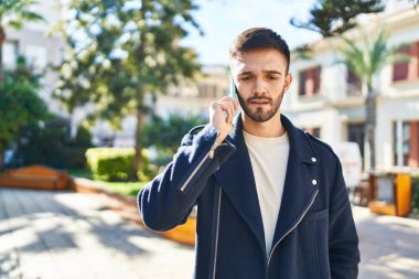 Young hispanic man talking on smartphone with serious expression at park