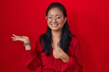 Asian young woman standing over red background showing palm hand and doing ok gesture with thumbs up, smiling happy and cheerful 