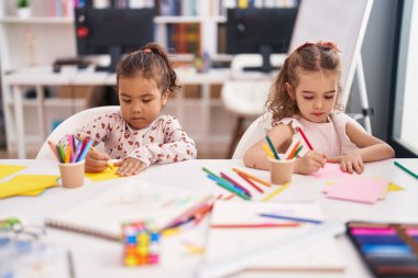 İki anaokulu öğrencisi sınıfta masaya oturmuş kağıda resim çiziyor.