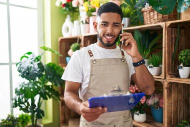 Young latin man florist talking on smartphone reading document at flower shop