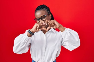 African woman with braids standing over red background smiling in love doing heart symbol shape with hands. romantic concept. 