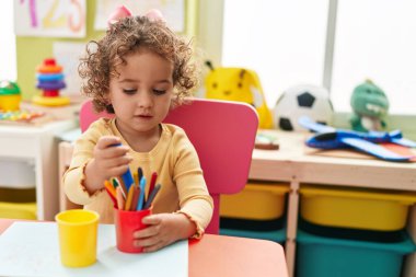 Adorable hispanic toddler student sitting on table drawing on paper at kindergarten