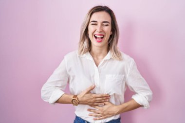 Young beautiful woman standing over pink background smiling and laughing hard out loud because funny crazy joke with hands on body. 