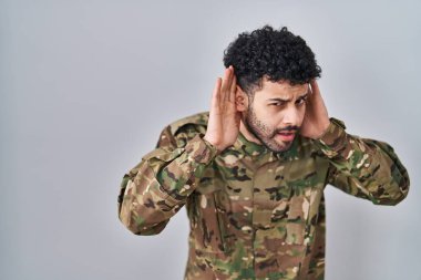 Arab man wearing camouflage army uniform trying to hear both hands on ear gesture, curious for gossip. hearing problem, deaf 