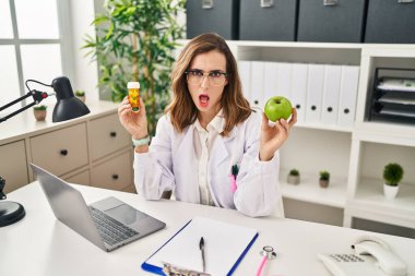 Young woman working at dietitian clinic in shock face, looking skeptical and sarcastic, surprised with open mouth 