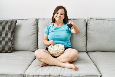 Down sendromlu esmer kadın kanepede oturup televizyon izliyor oturma odasında patlamış mısır yiyor.