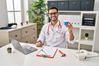 Young hispanic doctor man with beard holding ear cotton buds looking positive and happy standing and smiling with a confident smile showing teeth 
