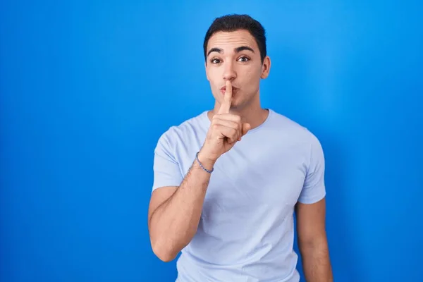 青い背景の上に立っている若いヒスパニック系の男が唇に指で静かにするように求めている 沈黙と秘密の概念 — ストック写真