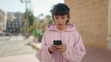 Young hispanic woman surprised using smartphone at street