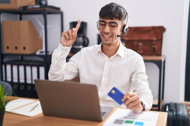 Young hispanic man working using computer laptop holding credit card smiling amazed and surprised and pointing up with fingers and raised arms. 