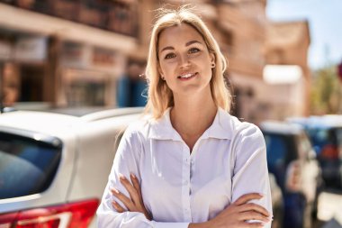 Genç sarışın kadın gülümsüyor. Kollarını kavuşturup caddede duruyor.