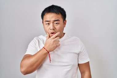 Young chinese man standing over white background looking fascinated with disbelief, surprise and amazed expression with hands on chin 