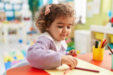 Adorable hispanic girl preschool student sitting on table drawing on paper at kindergarten