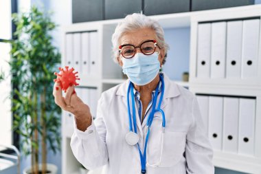 Doktor üniforması giyen yaşlı gri saçlı kadın ve klinikte elinde virüs oyuncağı tutan tıbbi maske.