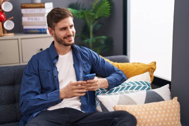 Beyaz bir adam akıllı telefon kullanıyor. Evdeki kanepede oturuyor.