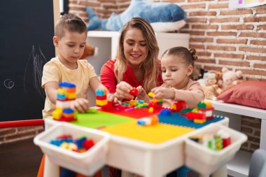 Teacher with boy and girl playing with construction blocks sitting on table at kindergarten