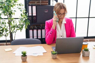 Young blonde girl business worker using laptop talking on smartphone at office