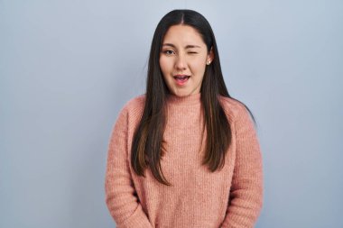 Young latin woman standing over blue background winking looking at the camera with sexy expression, cheerful and happy face. 