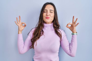 Mavi arka planda duran esmer kadın rahatlamış ve parmaklarıyla meditasyon hareketi yaparken gözleri kapalı gülümsüyor. yoga konsepti. 