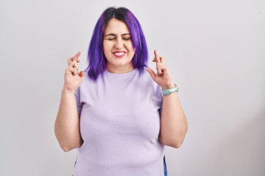 Plus size woman wit purple hair standing over isolated background gesturing finger crossed smiling with hope and eyes closed. luck and superstitious concept. 