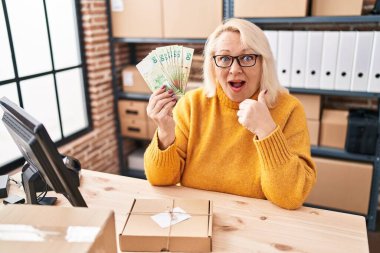 Middle age caucasian woman working at small business ecommerce holding hong kong dollars scared and amazed with open mouth for surprise, disbelief face 