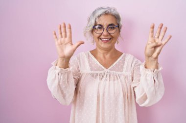 Pembe arka planda gri saçlı orta yaşlı bir kadın dokuz numaralı parmaklarıyla işaret ederken kendinden emin ve mutlu bir şekilde gülümsüyor.. 