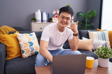 Young asian man using laptop at home sitting on the sofa smiling doing phone gesture with hand and fingers like talking on the telephone. communicating concepts. 