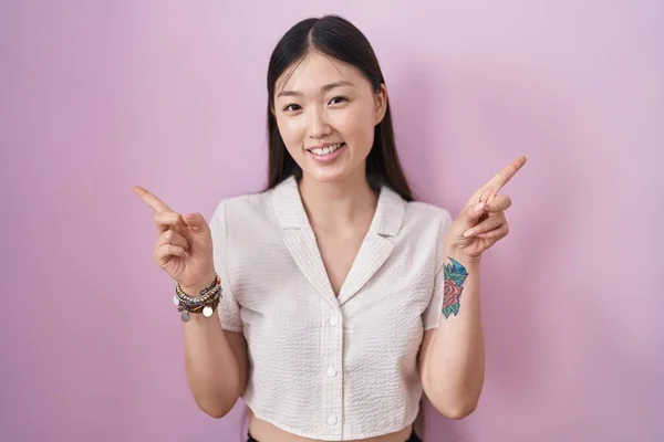 ピンクの背景に立っている中国の若い女性は さまざまな方向に指で自信を持って指して笑っている 広告のコピースペース — ストック写真