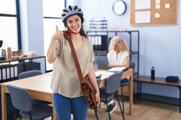 Young brunette woman working at the office wearing bike helmet smiling happy and positive, thumb up doing excellent and approval sign