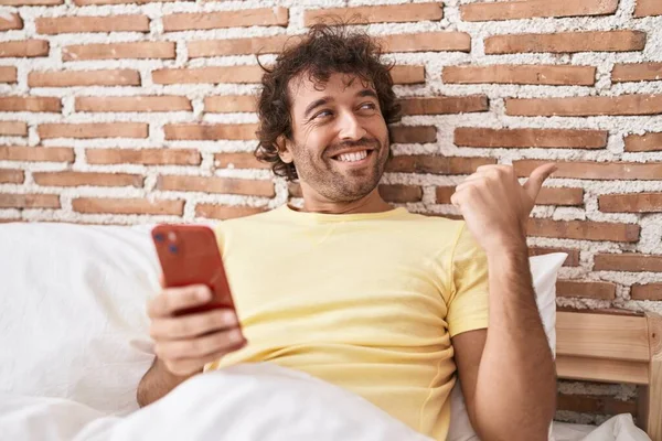 Hispanic Young Man Using Smartphone Bed Pointing Thumb Side Smiling — Stock Fotó
