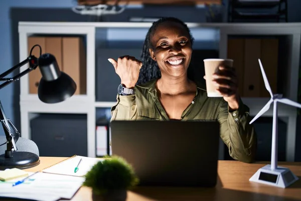 African woman working using computer laptop at night pointing to the back behind with hand and thumbs up, smiling confident