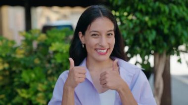 Young beautiful hispanic woman smiling confident doing ok sign with thumbs up at street
