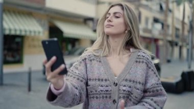 Young blonde woman smiling confident making selfie by the smartphone at street