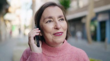 Orta yaşlı bir kadın gülümsüyor. Sokakta akıllı telefondan konuşuyor.