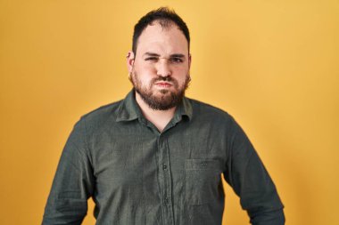 Plus size hispanic man with beard standing over yellow background puffing cheeks with funny face. mouth inflated with air, crazy expression. 