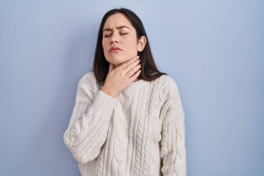 Genç esmer kadın mavi arka planda duruyor. Ağrılı boynuna dokunuyor. Grip, mantar ve enfeksiyon yüzünden boğazı ağrıyor. 