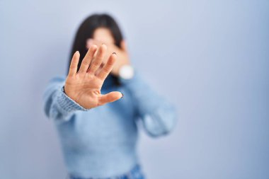 Mavi arka planda duran genç İspanyol kadın elleriyle gözlerini kapatıyor ve üzgün ve korku ifadesiyle dur hareketi yapıyor. utangaç ve olumsuz kavram. 