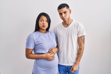 Young hispanic couple expecting a baby standing over background puffing cheeks with funny face. mouth inflated with air, crazy expression. 