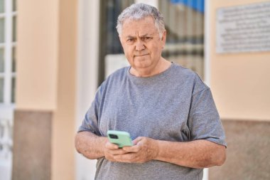 Orta yaşlı, kır saçlı bir adam sokakta rahat bir ifadeyle akıllı telefon kullanıyor.