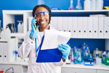 Bilim adamı üniforması giyen Afrikalı Amerikalı kadın akıllı telefon laboratuvarında konuşuyor.