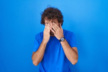 Hispanic young man standing over blue background rubbing eyes for fatigue and headache, sleepy and tired expression. vision problem 