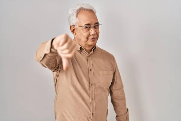 Hispanic senior man wearing glasses looking unhappy and angry showing rejection and negative with thumbs down gesture. bad expression.