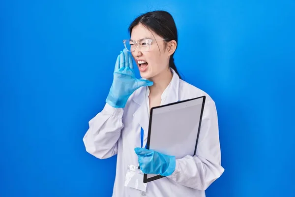 Chinese young woman working at scientist laboratory shouting and screaming loud to side with hand on mouth. communication concept.