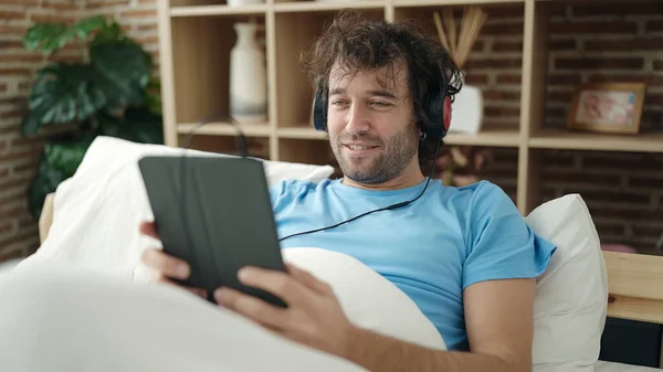 Young Hispanic Man Using Touchpad Headphones Lying Bed Bedroom — Zdjęcie stockowe