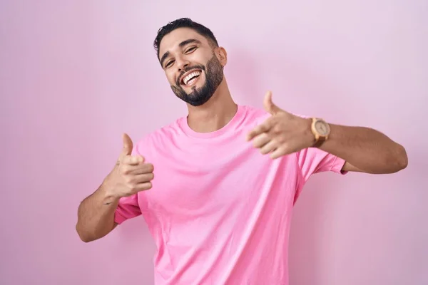 ヒスパニック系の若い男がピンクの背景に立って幸せと面白い顔を持つカメラに指を指しています 良いエネルギーと振動は — ストック写真