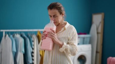 Middle age woman smiling confident smelling detergent bottle at laundry room