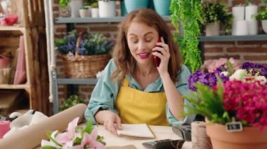 Young beautiful hispanic woman florist talking on smartphone reading notebook at florist