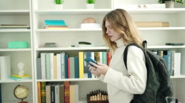Kütüphane üniversitesinde rafların olduğu kitabı tutan sarışın genç bir kız öğrenci.