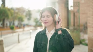 Genç beyaz kadın, kendine güvenen bir şekilde gülümsüyor müzik dinliyor ve sokakta dans ediyor.