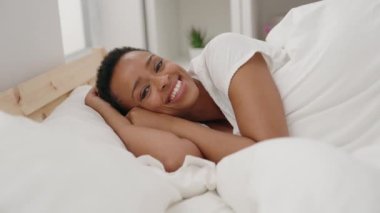 Afro-Amerikalı kadın, yatakta kendinden emin bir şekilde yatarken gülümsüyor.